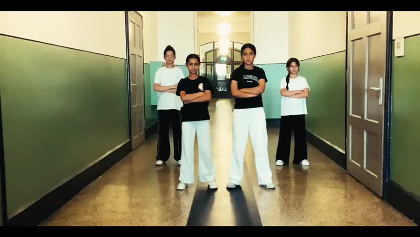 Vier junge Menschen stehen mit verschränkten Armen in einem Flur. Sie tragen lässige Sportkleidung und schauen mit ernster Miene in die Kamera. Der Flur hat grüne Wände und Türen auf jeder Seite.