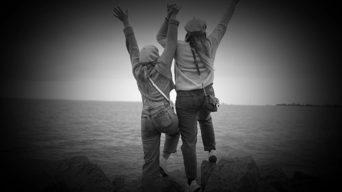 Zwei Personen stehen auf einem Felsen am Wasser, den Blick abgewandt und mit feierlich erhobenen Armen. Beide tragen Freizeitkleidung und Hüte. Das schwarz-weiße Bild mit Vignetteneffekt weckt Abenteuerlust.