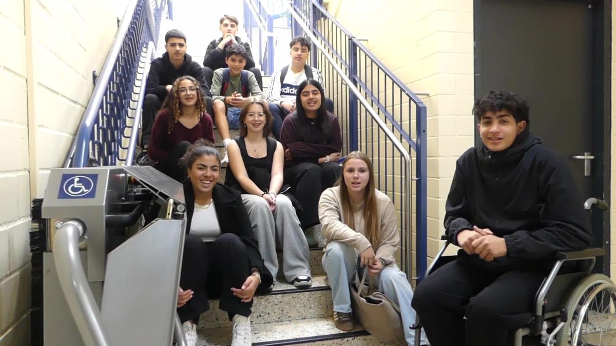 Eine Gruppe von Jugendlichen, darunter ein Rollstuhlfahrer, sitzt lächelnd auf einer Treppe neben einem barrierefreien Eingang.